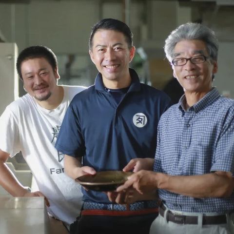 Three generations of Osada farmers standing together in what appears to be a pottery or ceramic workshop, with the central man holding a small round ceramic dish. The man on the left wears a white t-shirt, the center man wears a navy blue polo shirt with a white circular logo, and the man on the right wears a blue-and-white checkered button-up shirt.